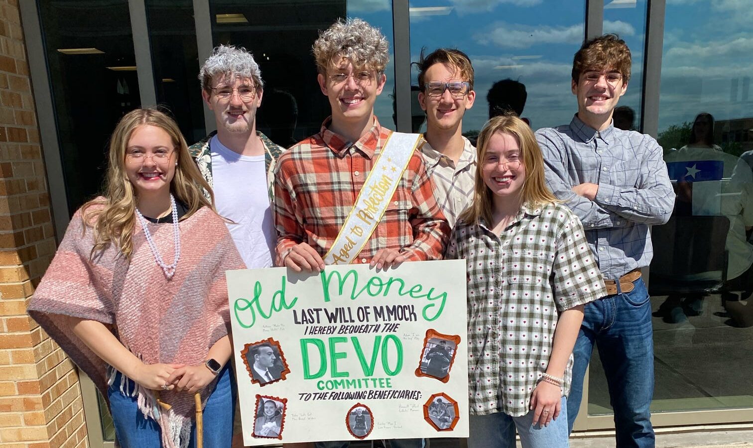 MSC ALOT's DEVO sub-committee dressed in "old money" themed attire, holding up a handmade poster about DEVO
